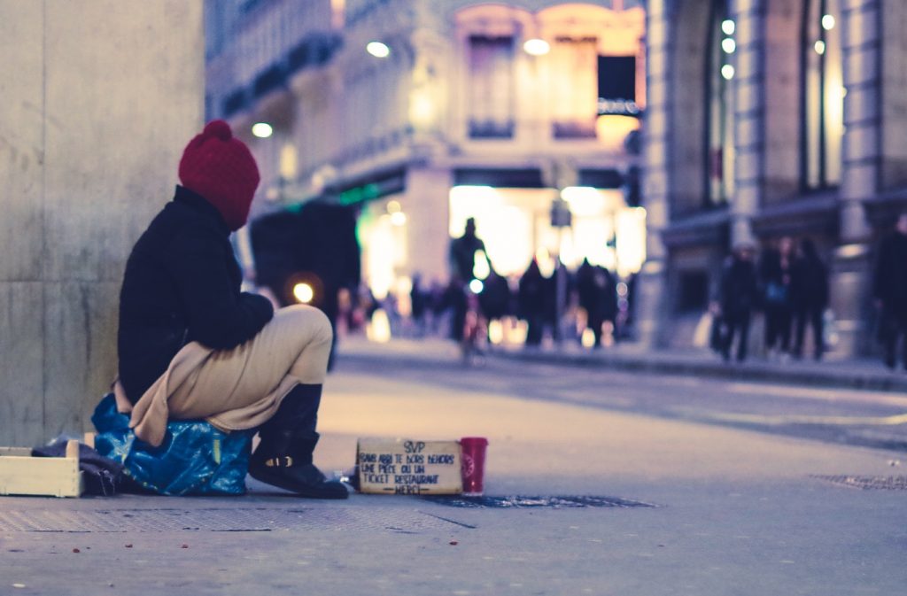 Obdachlose Person sitzt eingehüllt in eine Decke auf einem Platz in einer Stadt. Es ist Abend und vor ihr steht ein Pappbecher um Spenden zu sammeln.