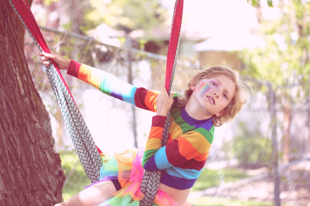 Ein Kind in Regenbogenpullover spielt an einen Baum auf einer bunten Hängematte. Im Gesicht ein Regenbogen.